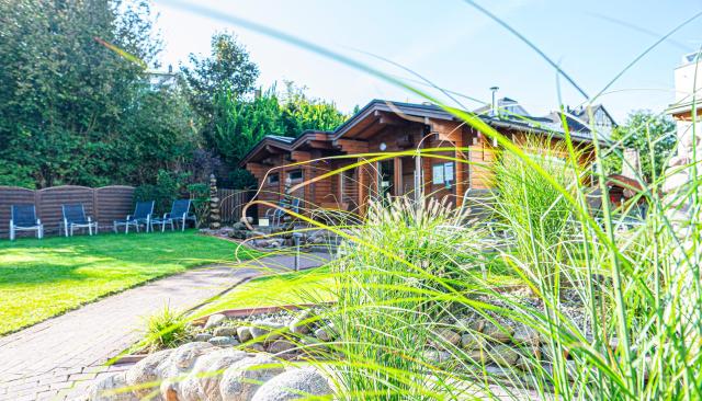 Eine sonnige Gartenanlage mit gepflegtem Rasen und Liegestühlen. Im Hintergrund stehen gemütliche Holzhäuser, die von grünen Büschen und Bäumen umgeben sind. Im Vordergrund rahmen hohe Gräser den Blick ein und verleihen der Szene eine natürliche, entspannte Atmosphäre.