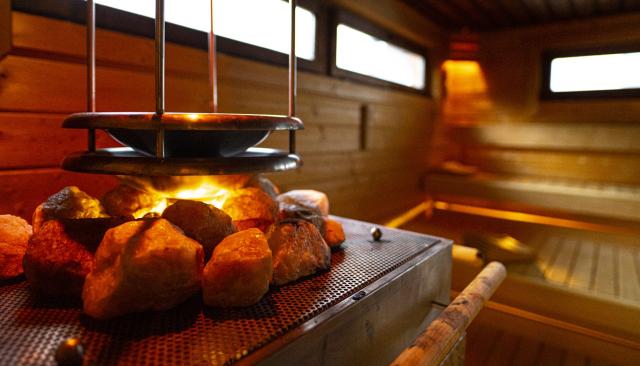 Ein warm erleuchteter Saunaraum mit hölzernen Bänken. Im Vordergrund glühen Saunasteine auf einem Ofen, aus denen sanftes Licht strahlt und eine behagliche Atmosphäre schafft.